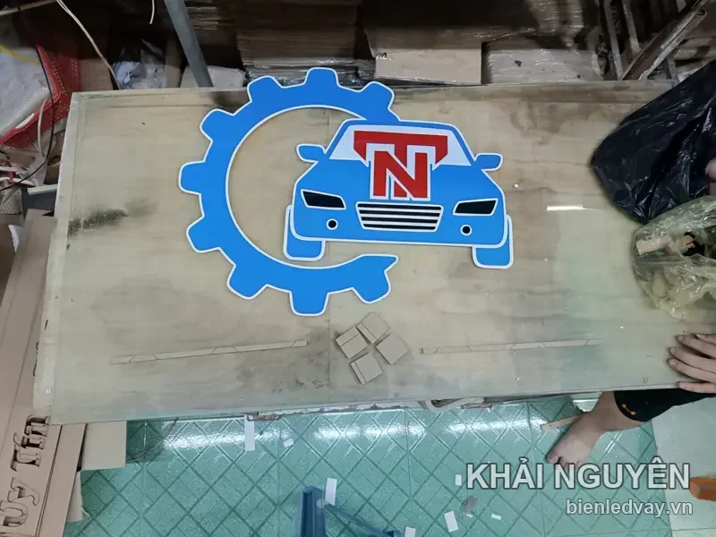 Logo mica cắt hình bánh răng xanh dương ôm chiếc ô tô, chữ N đỏ trên nóc xe, đặt trên tấm gỗ tại xưởng