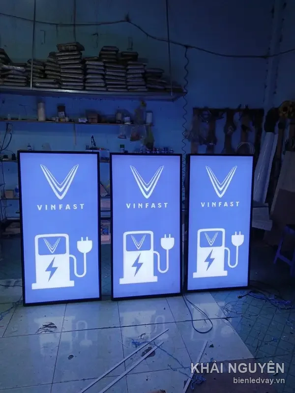 Ba hộp đèn VinFast chụp cận, logo chữ V và hình trụ sạc điện, kệ vật liệu xếp cao sát trần phía sau