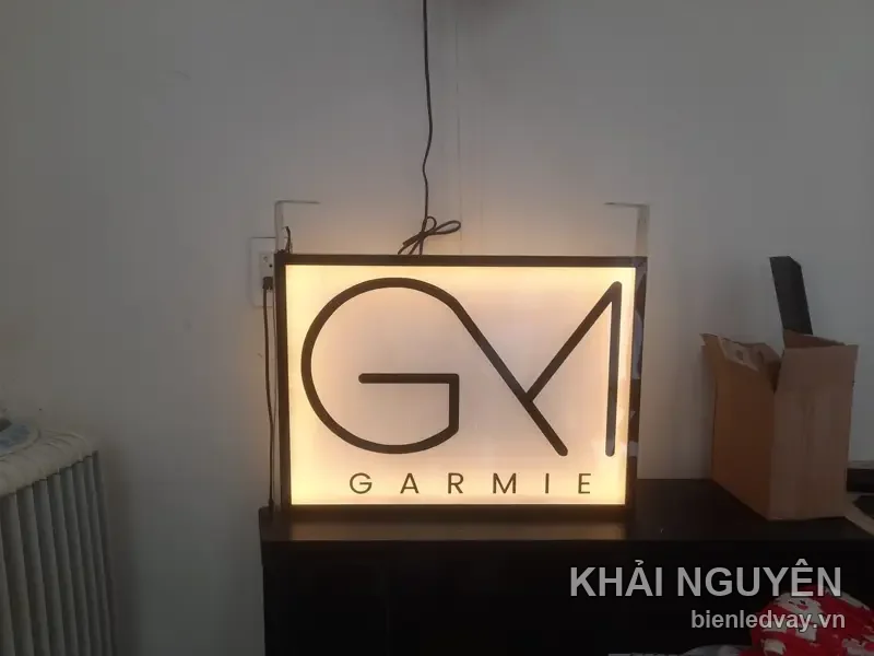 Hộp đèn GARMIE hắt sáng vàng ấm, logo GM nét mảnh màu nâu, kê trên tủ sẫm màu, dây điện thả từ trên xuống