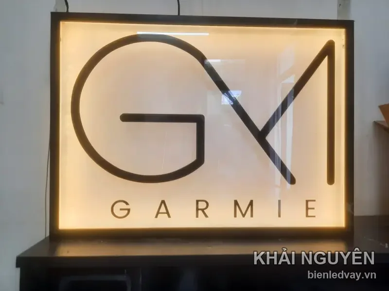 Cận cảnh hộp đèn GARMIE, logo GM lồng nét mảnh và chữ GARMIE trên nền mica sáng vàng, khung viền đen