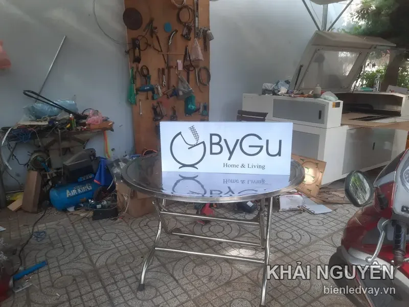 Bảng mica trắng in logo ByGu Home Living chữ đen, dựng trên bàn inox tròn giữa xưởng ngổn ngang máy móc