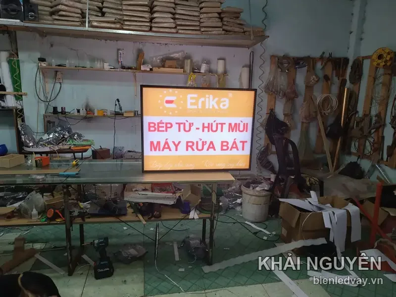 Hộp đèn Erika nền trắng, logo cam, chữ đỏ BẾP TỪ - HÚT MÙI, MÁY RỬA BÁT, mặt phẳng không viền LED, dựng trên bàn xưởng