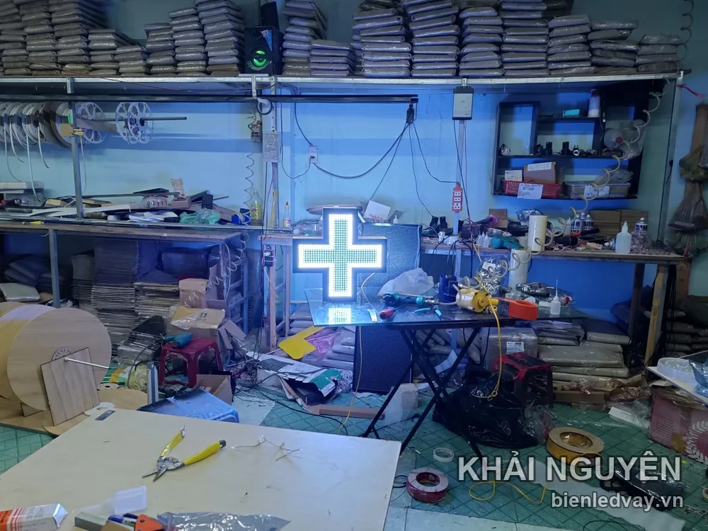 Đèn LED chữ thập nhà thuốc mặt ma trận trắng xanh, đặt trên bàn kính giữa xưởng ngổn ngang cuộn dây và vật tư