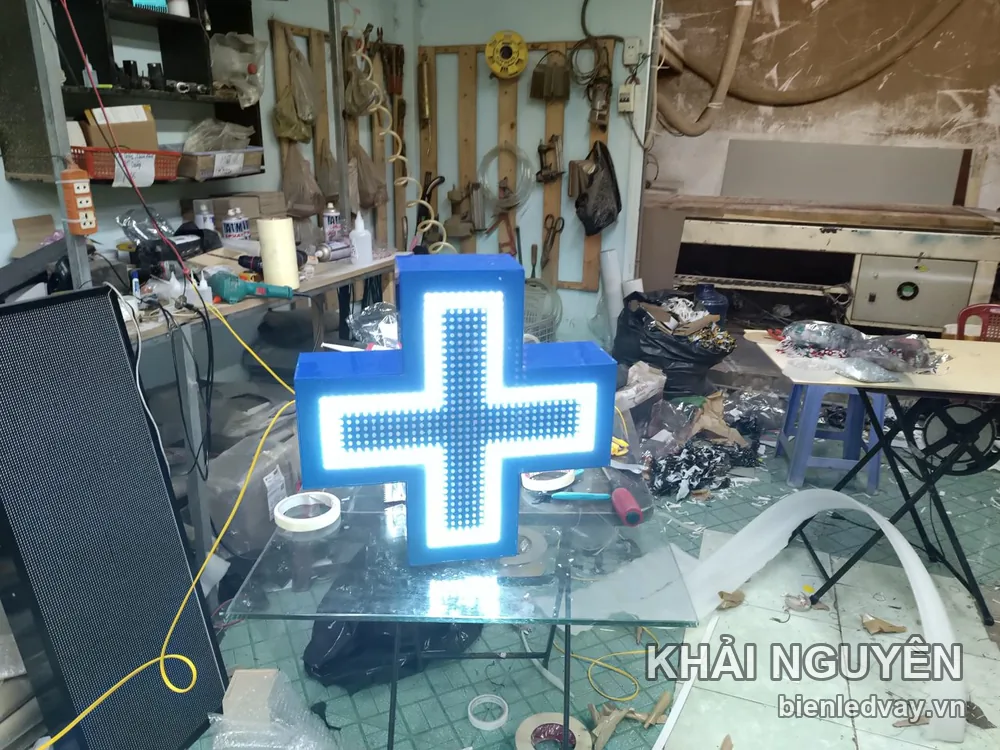 Đèn LED hình chữ thập vỏ nhựa xanh dương, mặt LED trắng sáng, dựng trên bàn kính trong xưởng cơ khí